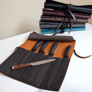 K&ouml;nnte beinhalten: Eine dunkelbraune Messertasche aus Leder mit beigefarbenem Innenfutter, die mehrere F&auml;cher f&uuml;r vier Messer mit schwarzen Griffen bietet, sowie ein weiteres Messer mit Holzgriff und silberner Klinge, das quer dar&uuml;ber liegt.