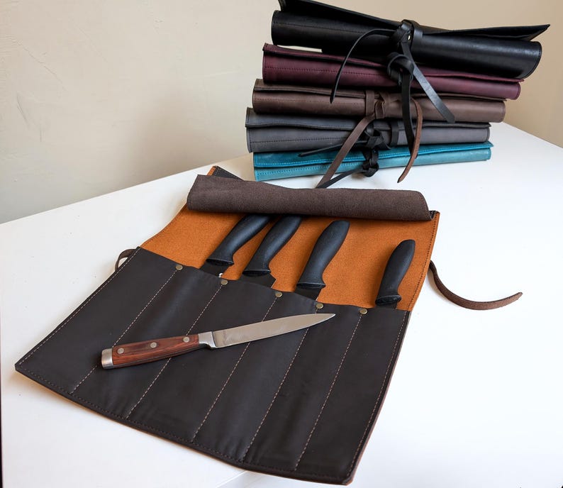 K&ouml;nnte beinhalten: Eine dunkelbraune Messertasche aus Leder mit beigefarbenem Innenfutter, die mehrere F&auml;cher f&uuml;r vier Messer mit schwarzen Griffen bietet, sowie ein weiteres Messer mit Holzgriff und silberner Klinge, das quer dar&uuml;ber liegt.