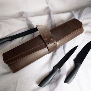 May include: A dark brown leather knife roll with a wrap-around strap secured by metal rivets. Three black kitchen knives are positioned around the roll, including one serrated blade and two smooth-edged blades, all featuring matte black handles.