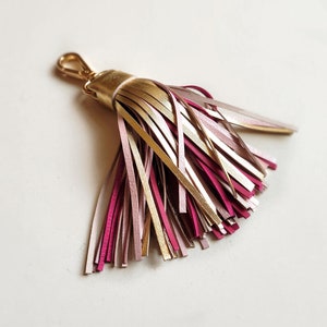 Peut inclure: Un porte-clés en forme de pompon doré et rose avec une boucle en métal doré. Le pompon est composé de fines lanières de cuir dans des tons de rose et d'or.