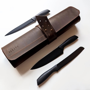 Puede incluir: Un estuche de cuchillos de cuero marrón con la palabra "GORDON" grabada en la parte delantera, acompañado de cuatro cuchillos negros. Los cuchillos incluyen un cuchillo de chef, un cuchillo dentado y otros dos cuchillos con mangos negros.