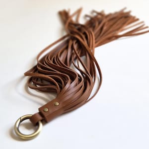 Op de afbeelding: Een bruine leren kwast met talloze lange, dunne stroken bevestigd aan een ronde metalen ring. De ring is met twee kleine, ronde metalen klinknagels aan de leren basis vastgemaakt, wat zorgt voor een functioneel accessoire met een strak ontwerp.