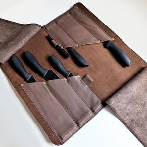 Könnte beinhalten: Eine braune Messertasche aus Leder mit mehreren Innentaschen, die mit Messingnieten befestigt sind. Die Tasche enthält vier Messer mit schwarzem Griff und einen schwarzen Stift sowie zwei große Seitenklappen zum Zusammenfalten.