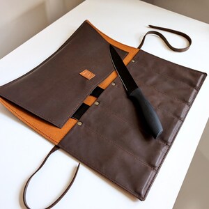 K&ouml;nnte beinhalten: Eine dunkelbraune Messertasche aus Leder mit beigefarbenem Innenfutter, ausgestattet mit mehreren vertikalen F&auml;chern, einem kleinen quadratischen Logo-Aufn&auml;her und einem langen Lederband zum Verschlie&szlig;en. Ein schwarzes K&uuml;chenmesser liegt auf der Tasche.