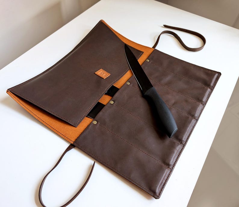 K&ouml;nnte beinhalten: Eine dunkelbraune Messertasche aus Leder mit beigefarbenem Innenfutter, ausgestattet mit mehreren vertikalen F&auml;chern, einem kleinen quadratischen Logo-Aufn&auml;her und einem langen Lederband zum Verschlie&szlig;en. Ein schwarzes K&uuml;chenmesser liegt auf der Tasche.