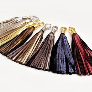 Könnte beinhalten: Eine Auswahl an Lederquasten in verschiedenen Farben, darunter Silber, Gold, Braun, Marineblau und Burgunderrot. Jede Quaste hat eine Metallkappe und einen Ring zur Befestigung. Die Quasten sind fächerförmig vor weißem Hintergrund angeordnet.