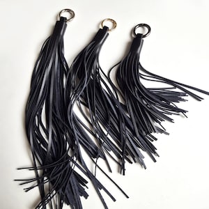 May include: Three black leather tassels of varying lengths, each featuring a smooth cylindrical top cap and a silver-toned metal ring attached to the top for hanging. The tassels consist of multiple thin, dangling strips of black leather.