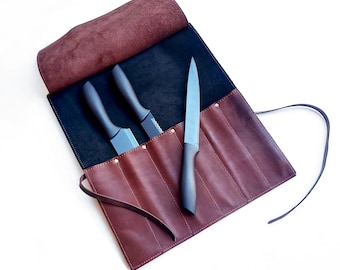 May include: A dark brown leather knife roll featuring a black interior lining with multiple slots. Two knives with black handles are tucked into the slots, while a third knife with a black handle and a metallic blade rests on the exterior.