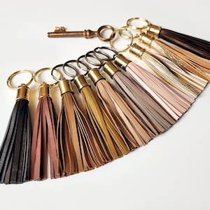 Könnte beinhalten: Eine Sammlung von Lederquasten-Schlüsselanhängern in verschiedenen Farben, darunter Schwarz, Braun, Gold und Beige. Jeder Schlüsselanhänger hat einen goldfarbenen Metallring und eine dekorative Quaste. Ein Schlüssel im Vintage-Stil ist ebenfalls vorhanden.