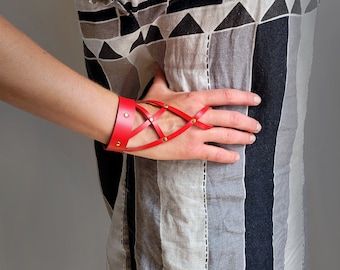 May include: A red leather bracelet with a criss-cross design and gold accents. The bracelet is worn on the wrist and extends to the fingers.