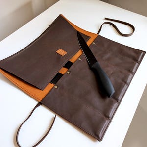 Könnte beinhalten: Eine dunkelbraune Messertasche aus Leder mit beigefarbenem Innenfutter, ausgestattet mit mehreren vertikalen Fächern, einem kleinen quadratischen Logo-Aufnäher und einem langen Lederband zum Verschließen. Ein schwarzes Küchenmesser liegt darin.