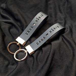 Puede incluir: Dos llaveros de cuero negro con herrajes dorados. Cada llavero tiene el nombre "DANA MARIS" grabado en relieve, con un símbolo de corazón entre los nombres. Los llaveros están sobre una tela negra.