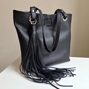 Pode incluir: Uma bolsa tote preta com superfície texturizada, apresentando duas alças de ombro presas com anéis de metal prateado. Uma longa borla de franjas pretas pende de um dos anéis. Um pequeno emblema quadrado preto com um logótipo está centrado.