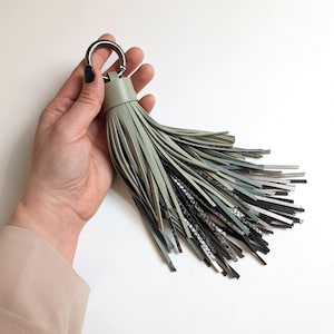 Könnte beinhalten: Ein großer Schlüsselanhänger mit Quaste aus salbeigrünem Leder und einem silbernen Metallring. Die Quaste weist eine Mischung aus Texturen und Farben auf, darunter schwarze und silberne Akzente, was ein stilvolles Accessoire ergibt.