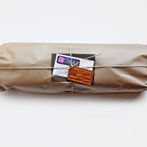 K&ouml;nnte beinhalten: Ein zylindrisches Paket, eingewickelt in braunes Kraftpapier und mit d&uuml;nner Schnur verschn&uuml;rt. An der Schnur befestigt ist ein wei&szlig;es rechteckiges Etikett mit dem Text ERIKAS sowie ein kleiner brauner Lederanh&auml;nger mit Thank You.