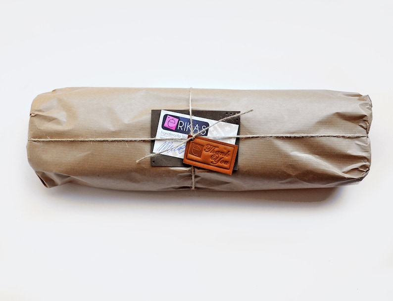 K&ouml;nnte beinhalten: Ein zylindrisches Paket, eingewickelt in braunes Kraftpapier und mit d&uuml;nner Schnur verschn&uuml;rt. An der Schnur befestigt ist ein wei&szlig;es rechteckiges Etikett mit dem Text ERIKAS sowie ein kleiner brauner Lederanh&auml;nger mit Thank You.