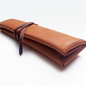 May include: A folded, light brown leather case with a dark brown leather strap wrapped around it. The case is closed with a leather tie. The edges of the case are stitched with a contrasting thread.