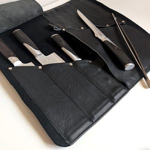 Könnte beinhalten: Eine schwarze Messertasche aus Leder mit mehreren Innentaschen, die drei Küchenmesser mit dunkel gemaserten Holzgriffen und silbernen Kropfen enthält, sowie eine lange, schmale Pinzette aus Edelstahl, die an der Seite liegt.