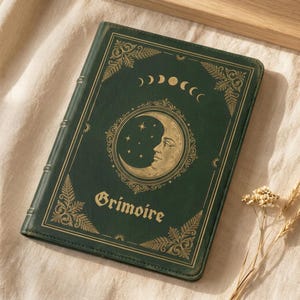 Könnte beinhalten: Ein dunkelgrünes Notizbuch mit Goldfolienprägung auf dem Einband. Das Design zeigt eine zentrale Mondsichel mit Gesicht, umgeben von Sternen, einem Mondphasenzyklus und Farnblättern in den Ecken. Das Wort Grimoire ist in goldenen Buchstaben aufgedruckt.