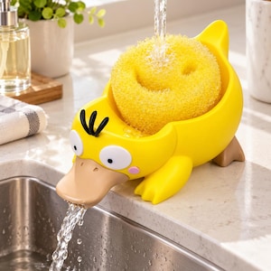 May include: A yellow duck-shaped sponge holder with a tan beak, white eyes, and black hair tufts. The holder contains a round yellow sponge with a smiley face cutout. Water flows from a faucet into the sponge and out through the duck's beak.