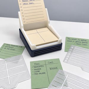 May include: A white and dark blue plastic note holder containing a stack of beige paper with a checklist. Surrounding the holder are three clear plastic grid templates and two green paper notes with handwritten text including lists and a quote.