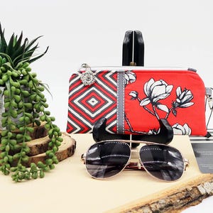 May include: A red and white floral print clutch with a silver zipper pull and a geometric pattern on the left side. A pair of sunglasses with gold frames and black lenses rests in front of the clutch. A small succulent plant is in the background.
