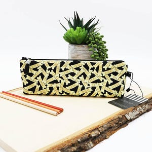 May include: Black pencil case with a repeating pattern of dog bones that say "TREAT" in gold. The pencil case has a black zipper and tassel. Three pencils are on a wooden board.