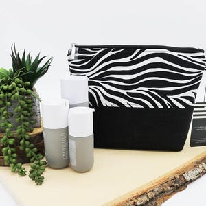 Può includere: Un beauty case con stampa zebrata in bianco e nero con base nera, accanto a tre flaconi grigi e bianchi. Nella foto sono presenti anche una piccola pianta in vaso e una lastra di legno.