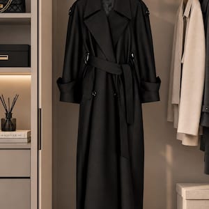 May include: A long black trench coat featuring wide lapels, a matching fabric belt tied at the waist, shoulder epaulets, and cuffed sleeves. The coat is hung on a wooden hanger inside a wardrobe, displaying a structured silhouette and smooth finish.
