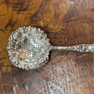 Könnte beinhalten: Ein Teesieb aus silberfarbenem Metall mit einem komplizierten geprägten Blumenmuster um die kreisförmige Schale und entlang des Griffs. Die Mitte der Schale enthält ein kleeblattförmiges perforiertes Design zum Abseihen von Teeblättern.