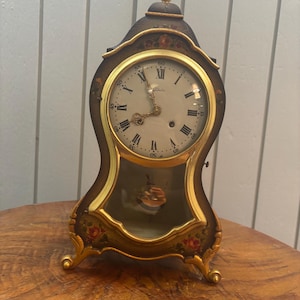 May include: A dark brown mantel clock with a gold-toned frame and finial. The clock face features black Roman numerals, ornate gold hands, and floral painted accents. A glass panel reveals a gold pendulum, and the base has four gold feet.