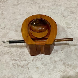 May include: A vintage wooden inkwell with a pen. The inkwell has a horseshoe-shaped design with a rounded top and a dark brown base. The pen is black with a silver nib. The inkwell is made of light and dark brown wood.