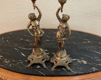 May include: Two antique bronze candle holders. Each features a classical female figure holding a floral torch. The figures stand on ornate bases. The candle holders are displayed on a black marble-topped table.
