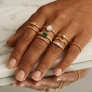 Puede incluir: Una colección de anillos dorados usados en los dedos, con varios diseños que incluyen un corazón pequeño, una piedra ovalada tipo ópalo, una piedra rectangular verde, una banda ondulada, una piedra triangular, una banda pavé y una banda trenzada.