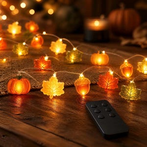May include: A string of LED fairy lights featuring translucent orange pumpkins, yellow and orange maple leaves, and brown acorns. The decorative lights are illuminated and include a small black rectangular remote control with six circular buttons.
