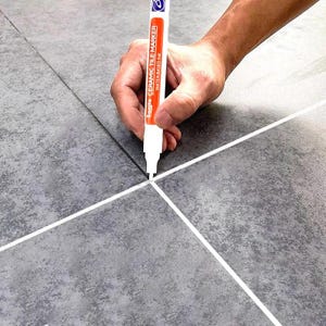 Peut inclure: Un marqueur pour carrelage blanc avec une étiquette orange portant le texte 'CERAMIC TILE MARKER WATER-BASED INK' utilisé pour appliquer une ligne blanche entre des carreaux de sol gris foncé texturés à l'aspect pierre.