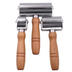 May include: A set of three hand rollers featuring polished silver metal cylinders and light brown wooden handles. Each tool consists of a cylindrical head attached to a metal frame, connected to a smooth, cylindrical wooden grip.