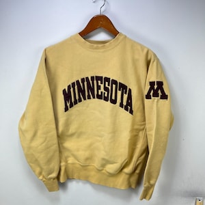 May include: A pale yellow crewneck sweatshirt featuring the word MINNESOTA in a dark maroon arched font across the chest. The right sleeve displays a dark maroon block letter M logo. The garment has ribbed cuffs and a ribbed hemline.