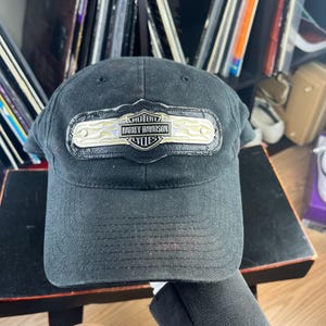 May include: A black baseball cap featuring a central rectangular patch with a metallic silver and gold finish. The patch displays the text Harley-Davidson Motor Cycles and includes a flame pattern design on the left and right sides of the logo.