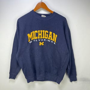 May include: A navy blue crewneck sweatshirt featuring the word MICHIGAN in large yellow letters arched across the chest, with the word WOLVERINES and a yellow block M logo printed in white and yellow underneath the main text.