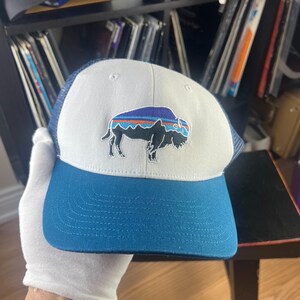 May include: A trucker cap featuring a white front panel, a dark blue brim, and a dark blue mesh back. The front panel displays an embroidered graphic of a bison with a stylised landscape design in shades of blue, orange, and black on its back.
