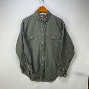 Könnte beinhalten: Ein langärmeliges Button-Down-Hemd in gedämpftem Olivgrün. Das Hemd verfügt über einen Button-Down-Kragen, zwei Brusttaschen mit geknöpften Klappen und ein kleines Tommy Hilfiger Logo-Etikett an der linken Tasche. Es hat eine durchgehende Knopfleiste.
