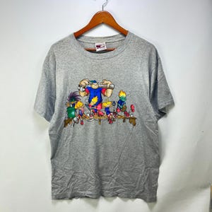 May include: A heather grey short-sleeve t-shirt featuring a colourful graphic print of five cartoon characters from the show Codename: Kids Next Door. The characters are depicted standing on a wooden platform, showcasing various outfits and hairstyles.