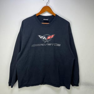 May include: A black long-sleeve shirt featuring a white, red, and black Corvette logo graphic on the chest. The logo includes a checkered flag and a red emblem inside a circle, with the word CORVETTE printed in white capital letters below.