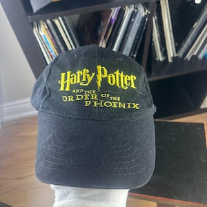 Puede incluir: Una gorra de béisbol negra con texto bordado en amarillo que dice Harry Potter and the Order of the Phoenix. La gorra tiene una corona estructurada y una visera curva con detalles de costura visibles en el panel frontal de la prenda.