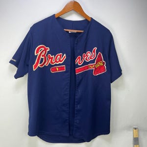 May include: A navy blue short-sleeved baseball jersey featuring the word Braves in red script with a yellow outline and a red tomahawk graphic. The Majestic logo is embroidered on the right sleeve. The jersey has a button-down front closure.