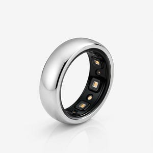 Peut inclure: Une bague connectée argentée avec un extérieur métallique poli et une doublure intérieure noire. La bande intérieure présente des capteurs électroniques intégrés, incluant de petits composants lumineux carrés et des points de contact circulaires.
