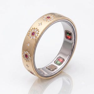 Peut inclure: Une bague connectée dorée avec une surface extérieure brossée ornée de motifs en étoile, chacun centré par une petite pierre rouge entourée de pierres claires. L'intérieur de l'anneau est argenté avec des capteurs électroniques visibles.
