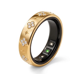 Puede incluir: Un anillo inteligente de tono dorado con un exterior pulido que presenta motivos de estrellas grabados y formas de flores de cuatro pétalos incrustadas con piedras claras. El interior de la banda es negro con sensores y una luz verde.