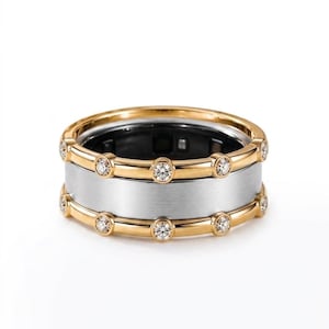 Peut inclure: Une bague large composée d'une section centrale en argent brossé encadrée par deux rails en or poli. Chaque rail doré est orné d'une série de petites pierres rondes et transparentes à facettes, réparties uniformément sur tout le pourtour.
